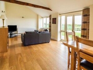 A living room with a sofa, a TV, wooden flooring, a dining table with chairs, and large windows with a view. - The Cart Shed Retreat in York (York)