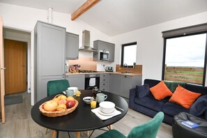 Ett modernt kök med grå skåp, en mikrovågsugn och en spis. Det finns ett matbord med frukt och en kaffebryggare. I vardagsrummet står en mörkblå soffa med orange kuddar, och ett fönster ger utsikt över landskapet.