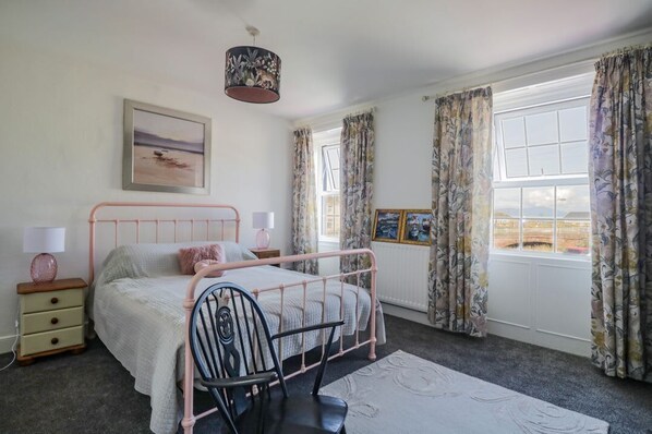 A bedroom with a bed, a chair, a nightstand, a lamp, a picture frame, and a window with curtains.
