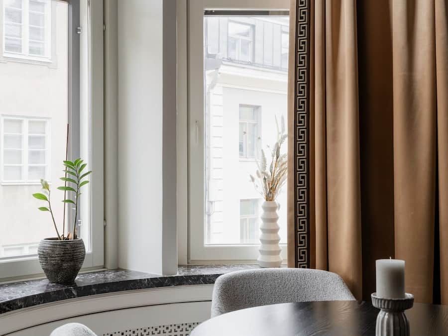 Um quarto com uma mesa redonda, duas cadeiras, um vaso de planta, uma vela e vista pela janela grande com cortina estampada.