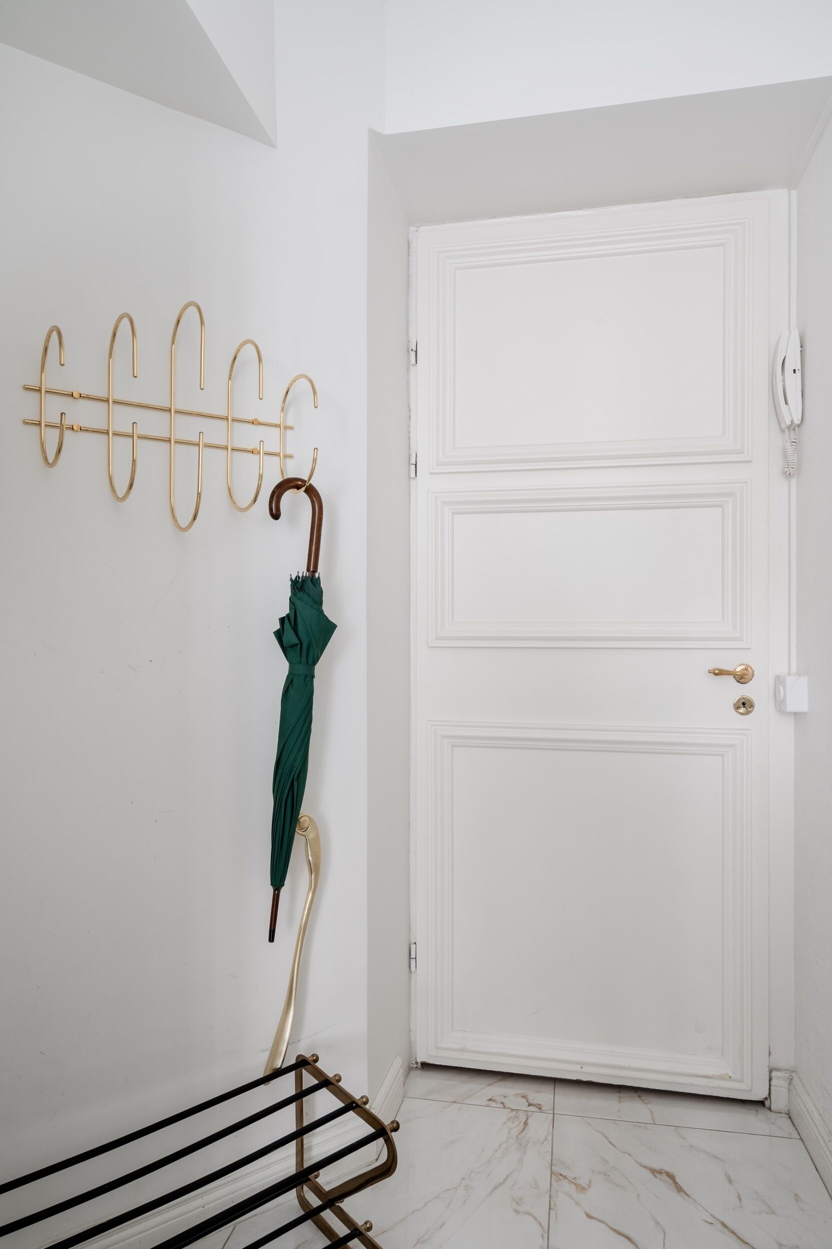 Uma porta branca com puxador de latão, um porta-roupas na parede com um guarda-chuva verde e um varal de metal preto.