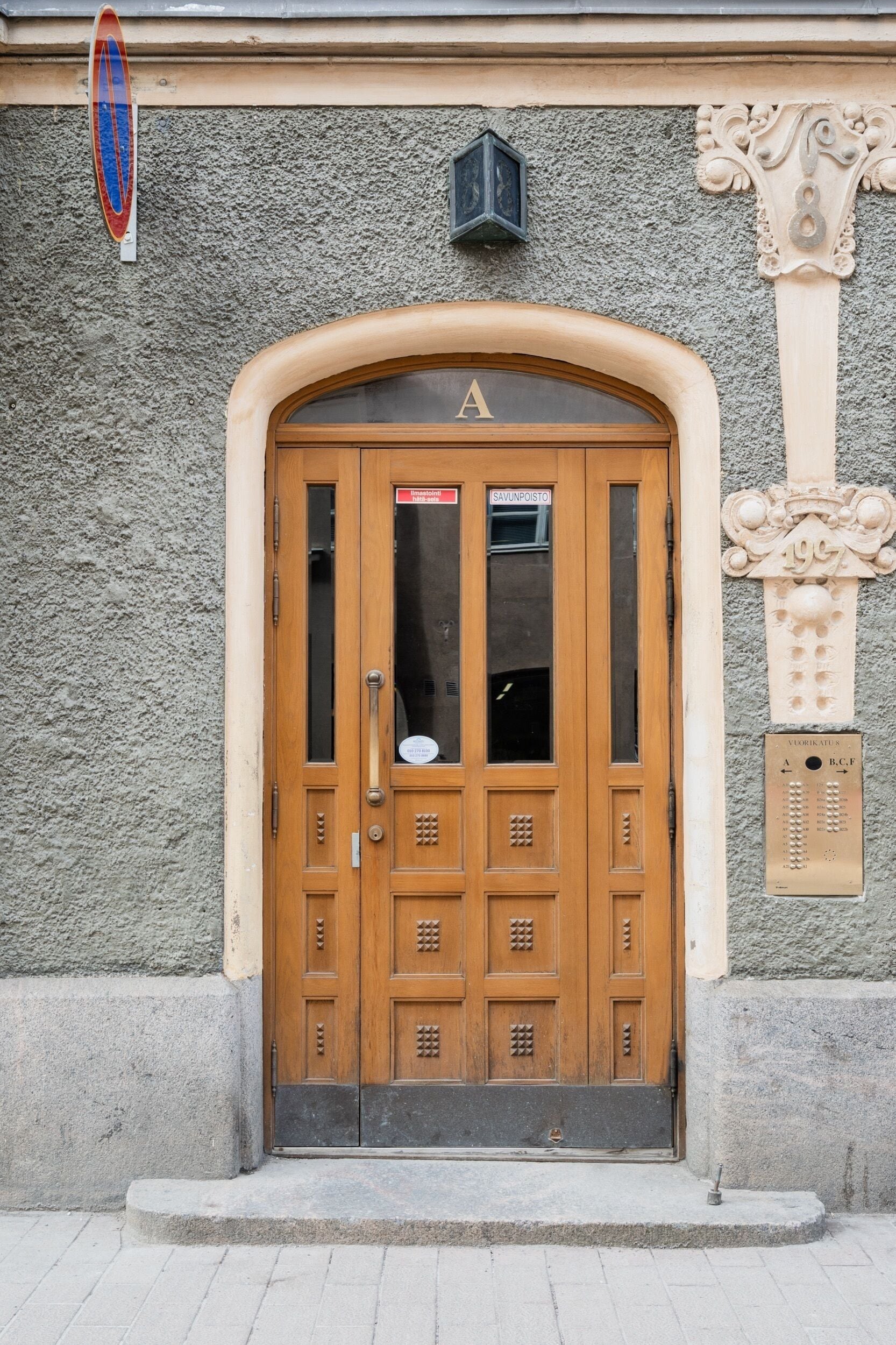 A wooden double door with a glass panel, a nameplate above the door, and a stone wall with a decorative column to the right.