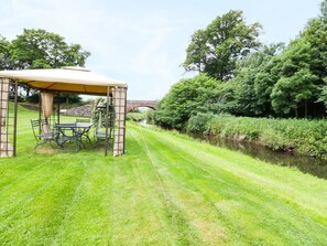 Ein Pavillon mit Tisch und Stühlen, ein Fluss und eine Brücke im Hintergrund.