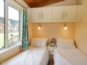 A bedroom with two beds, a window with curtains, a fan, and a small table with a book.