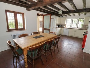 A dining area with a wooden table and chairs, a kitchen in the background, and a window with a view.