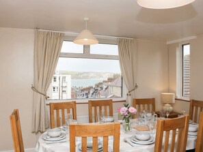 A dining area with a table set for six, wooden chairs, a vase with flowers, and a view of buildings and water through large windows.