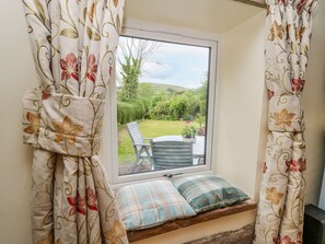 A window seat with a bench and cushions, curtains with floral patterns, and a view of a garden and outdoor furniture. - Buzzards Breg, Builth Wells (Builth Wells)