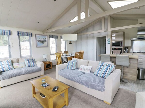 A spacious living area with two sofas, a coffee table, a dining table with chairs, a kitchen area with modern appliances, and large windows with striped curtains.