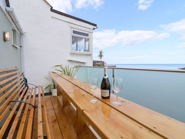 Un banco y una mesa de madera con una botella de Prosecco y dos copas en un balcón con vistas al mar tranquilo.