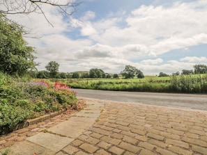 A paved pathway leading to a rural road with greenery and trees in the background.