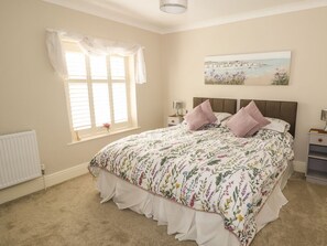 A bedroom with a large bed, a window with blinds, a bedside table with a lamp, and a painting on the wall.