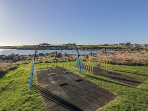 Ein Spielplatz mit Schaukeln und einer Rutsche, eine Rasenfläche und ein Gewässer im Hintergrund.