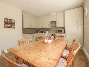 Un coin repas avec une table en bois, des chaises, un saladier de fruits et, au fond, une cuisine.