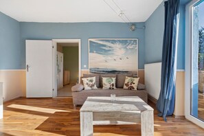 A living room with a wooden floor, a sofa with floral cushions, a painting of birds, and a view through a window.