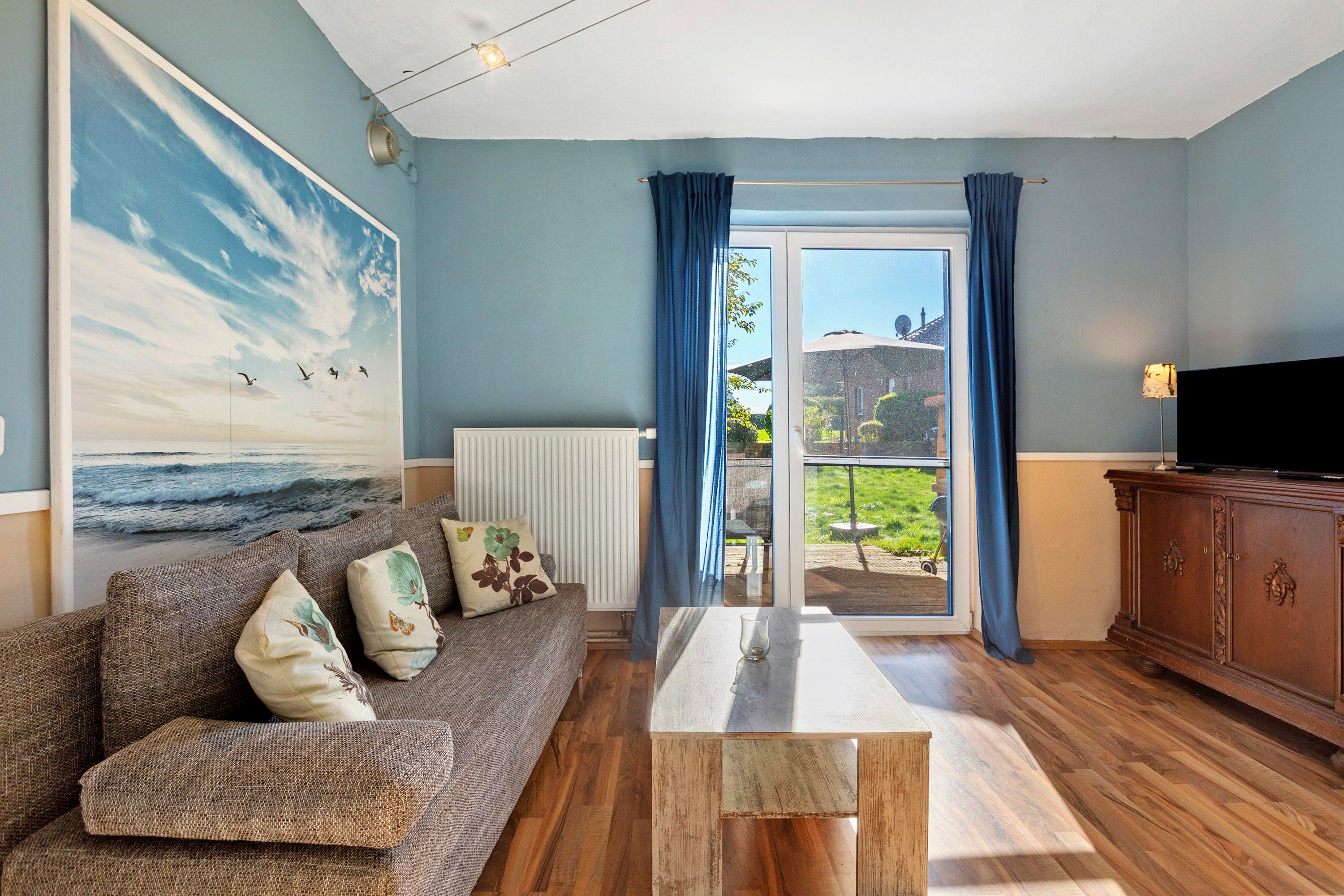 Ein Wohnzimmer mit einer Couch, einem Couchtisch, einem Seestück und einem Fernseher, mit Blick durch eine Glastür.