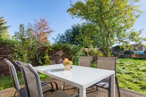 An outdoor patio with a white table and chairs, surrounded by greenery and a wooden fence.