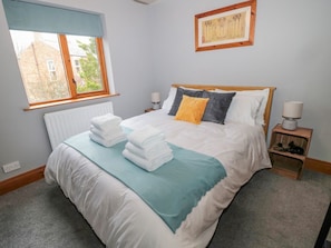 Cama bem arrumada com roupa de cama branca e verde-água, uma janela com persiana azul, um quadro na parede e uma pequena mesa de cabeceira com abajur.