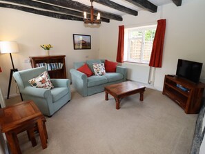 A living room with two light blue sofas, a wooden coffee table, a bookshelf, a television stand, and a window with curtains. - Mortons Cottage, Castlemorton (Malvern)