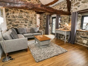 A cozy living room with a stone wall, wooden beams, a grey sofa, a wooden coffee table, and a stone fireplace. - Beudy Bach, Llanfair (Llanbedr)