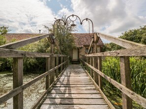 Eine Holzbrücke führt zu einem Gebäude mit einem Ziegeldach, umgeben von Grünpflanzen.