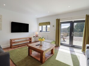 A modern living room with a flat-screen TV, a wooden coffee table with flowers, a bench, and a view through a glass door to an outdoor area. - The Old Dairy, Adiham, near Wingham, Kent. (Canterbury)