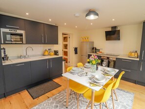 A modern kitchen with dark cabinets, a white countertop, a built-in oven, a refrigerator, and a dining area with a white table and yellow chairs. - Holly Cottage, Stratford-Upon-Avon (Stratford-upon-Avon)