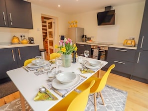 A dining area with a table set for four, a vase of flowers, and kitchen appliances in the background. - Holly Cottage, Stratford-Upon-Avon (Stratford-upon-Avon)