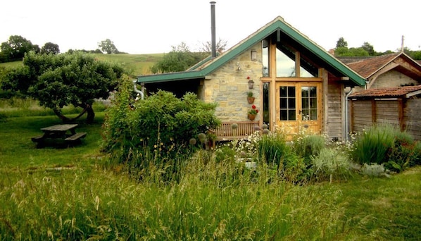Una casa de piedra rústica con techo verde, un porche de madera y una mesa de picnic en un campo cubierto de hierba.