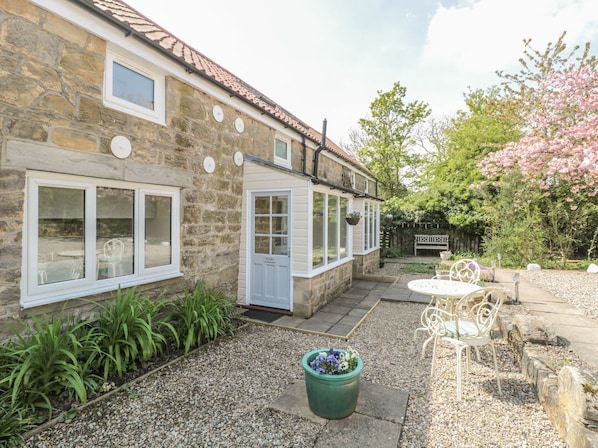 A stone building with a white door, a patio with a table and chairs, and a garden with plants and a tree. - Robin Cottage, Morpeth (Morpeth)