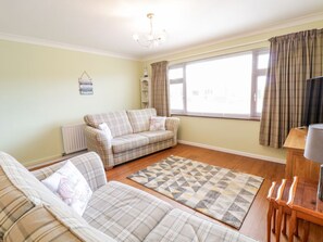 A living room with a sofa set, a patterned rug, a wooden table, and a window with curtains. - Rhos Deiniol, Morfa Bychan (Porthmadog)