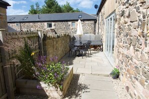 A stone building with a patio area featuring a table and chairs, surrounded by a wooden fence and flowering plants. - The Coach House, Bamburgh (Bamburgh)