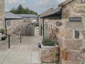 A stone building labeled 'The Coach House' with a wooden planter and a staircase leading to a covered outdoor seating area. - The Coach House, Bamburgh (Bamburgh)