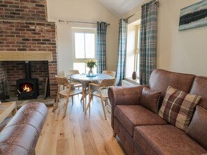A cozy living room with a fireplace, a brown leather sofa, a wooden dining table with chairs, and plaid curtains.