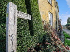 Ein hölzernes Wegweiser mit Beschilderung zum Cotswold Way und zum Broadway Tower, vor einer Kulisse aus einem efeuüberwucherten Gebäude und einem gepflegten Garten.