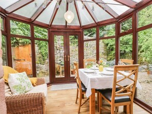 A conservatory with a wooden table set for dining, a wicker sofa, and a stone wall visible through the glass doors. - The Lodge, Greystoke (Penrith)