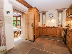 A rustic kitchen with wooden cabinets, a stone floor, and a dining area visible through an open door. - The Lodge, Greystoke (Penrith)