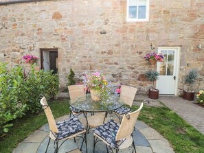 Un bâtiment en pierre avec une table en verre et des chaises en osier, entouré de plantes en pot et d’un petit jardin.