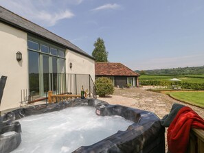 A hot tub in a courtyard with a building, a tree, and a view of the countryside. - The Hayloft, Honiton (Honiton)