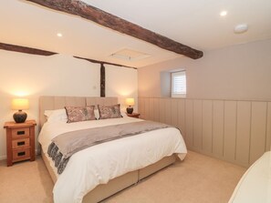A bedroom with a large bed, wooden headboard, two bedside tables with lamps, a window with blinds, and a sloped ceiling with exposed beams. - The Hayloft, Honiton (Honiton)