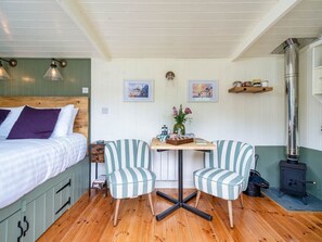 A cozy room with a bed, a table with chairs, a small table with decorative items, a wall-mounted shelf, and a wood-burning stove. - Onnen, near to Aberaeron. 1bed (Llanarth)