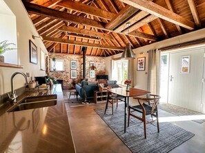 A rustic kitchen with a wooden ceiling, a dining area with a table and chairs, and a stone wall.