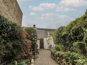 A narrow brick pathway leading to a white building with a wooden door, surrounded by lush greenery and potted plants. - Victoria Cottage, Amble (Amble)