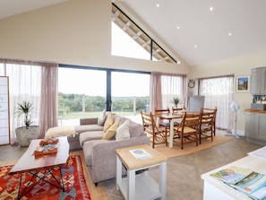 Ein großzügiger Wohnbereich mit Sofa, Couchtisch, Esstisch und Stühlen. Große Fenster bieten einen Ausblick, ein Deckenventilator sorgt für Luftzirkulation, und es gibt eine Küchenzeile.