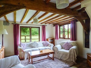 Ein gemütliches Zimmer mit Holzbalken, zwei Sofas, einem Tisch und großen Fenstern mit Vorhängen.
