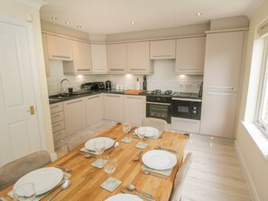 A modern kitchen with white cabinets, a wooden dining table set for four, and a window providing natural light. - The Shipwrights, Whitby (Whitby)