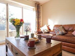A living room with a wooden coffee table, a brown leather sofa, and a window with curtains. - Farmer's Rest, Cambridgeshire. (Ely)