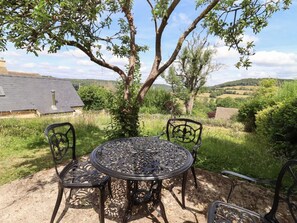 Un espacio al aire libre con una mesa y sillas metálicas redondas, rodeado de vegetación y vistas a un paisaje rural.