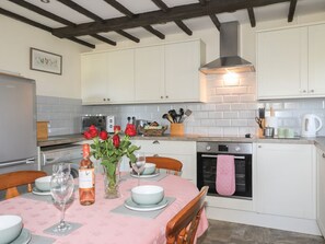A kitchen with white cabinets, a brick backsplash, a gas stove, and a dining table set with plates, glasses, and a vase of red roses. - Pabo Lodge: A Cozy Retreat (Llandudno Junction)