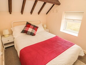 A bed with white bedding, a red blanket, and a checkered pillow. There is a nightstand with a lamp, a window with blinds, and a wooden ceiling beam.