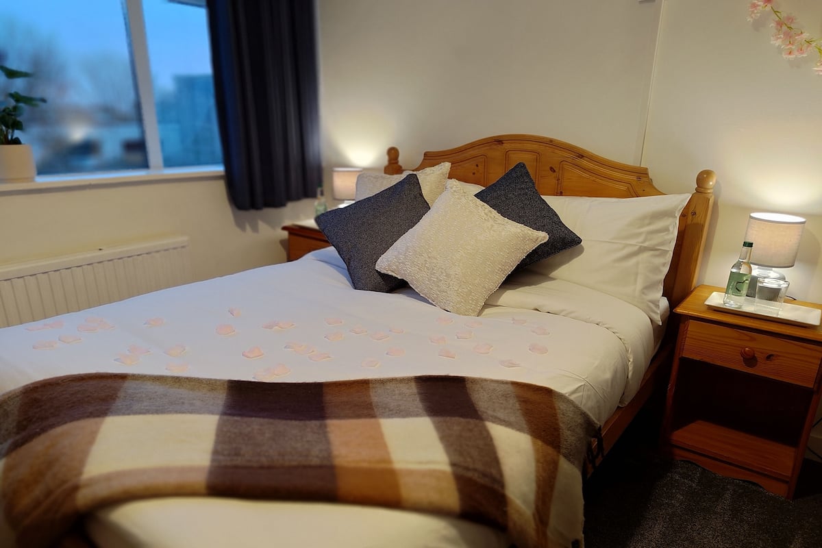 A neatly made bed with a checkered blanket, a bedside table with a lamp, and a window with curtains.